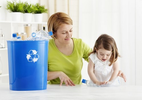 A woman with short, light brown hair wearing a lime green top is sitting at a table indoors, assisting a young girl with shoulder-length brown hair in a white top, as they handle clear plastic bottles. The bottles are partially crushed and appear to be empty. To the left, there is a large blue recycling bin with a white recycling symbol, filled with more empty plastic bottles. The background features a bright, modern kitchen with white shelves holding potted plants and various kitchen items, suggesting a clean, organized environment. The scene appears to promote eco-friendly waste disposal practices, consistent with the services offered by House Clearance Stevenage, a company specializing in rubbish removal within the local area, including nearby Stevenage and surrounding postcode areas. The natural lighting emphasizes the practical aspect of recycling and waste sorting, fitting within the context of domestic rubbish management and house clearance services in Hertfordshire.
