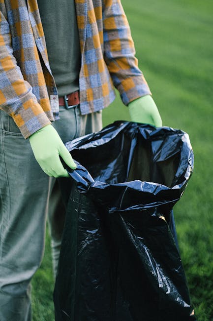 Three large black plastic rubbish bags are placed on a paved sidewalk in front of a black metal fence, with dense foliage visible behind the fence and a building partially visible to the right. The ba