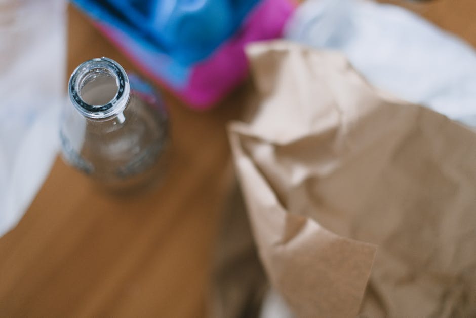 A clear glass bottle with a narrow neck and open top sits on a wooden surface, surrounded by crumpled brown paper bags and blurred colorful packaging in the background. The bottle appears to be empty 