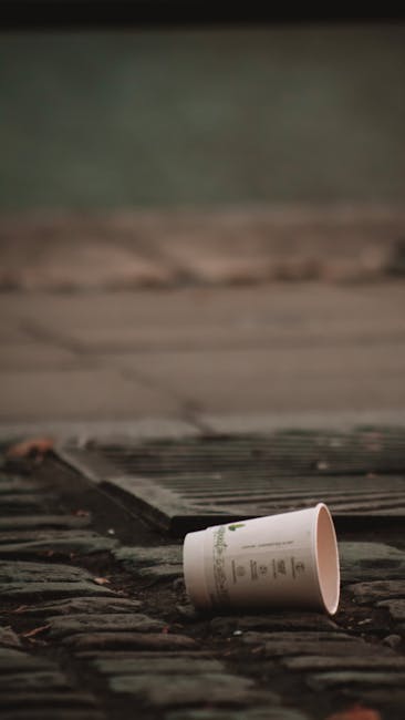 A clear glass bottle with a narrow neck and open top sits on a wooden surface, surrounded by crumpled brown paper bags and blurred colorful packaging in the background. The bottle appears to be empty 