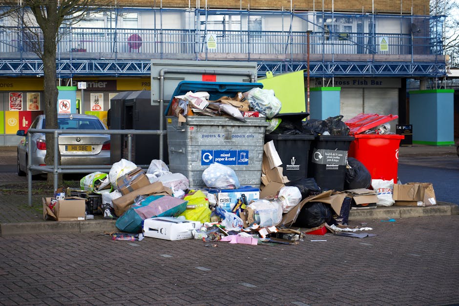 A small blue cargo truck parked parallel to a curb on a street, loaded with various types of refuse including large cardboard boxes, black plastic garbage bags, wooden pallets, and miscellaneous house