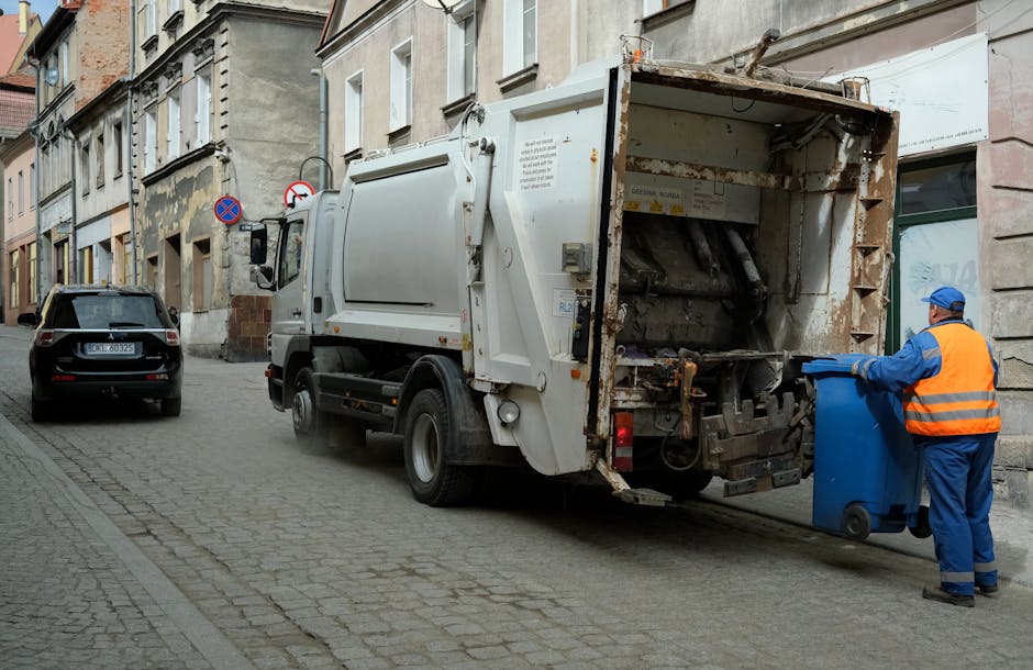 A collection of overflowing rubbish bins and scattered waste materials on a paved urban sidewalk area. The scene includes a large grey mixed waste bin with a blue lid, positioned centrally, filled wit
